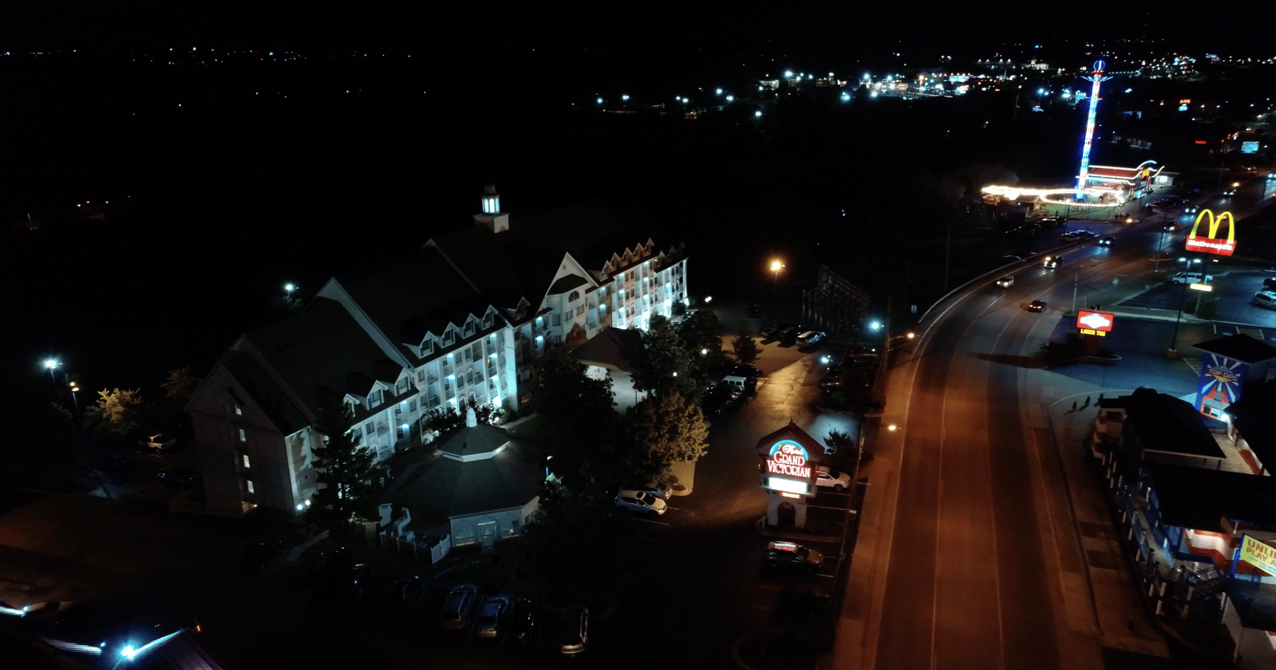 A nighttime aerial view of a multi-story white building with a dark roof, lit up at night, with a parking lot and trees in front.